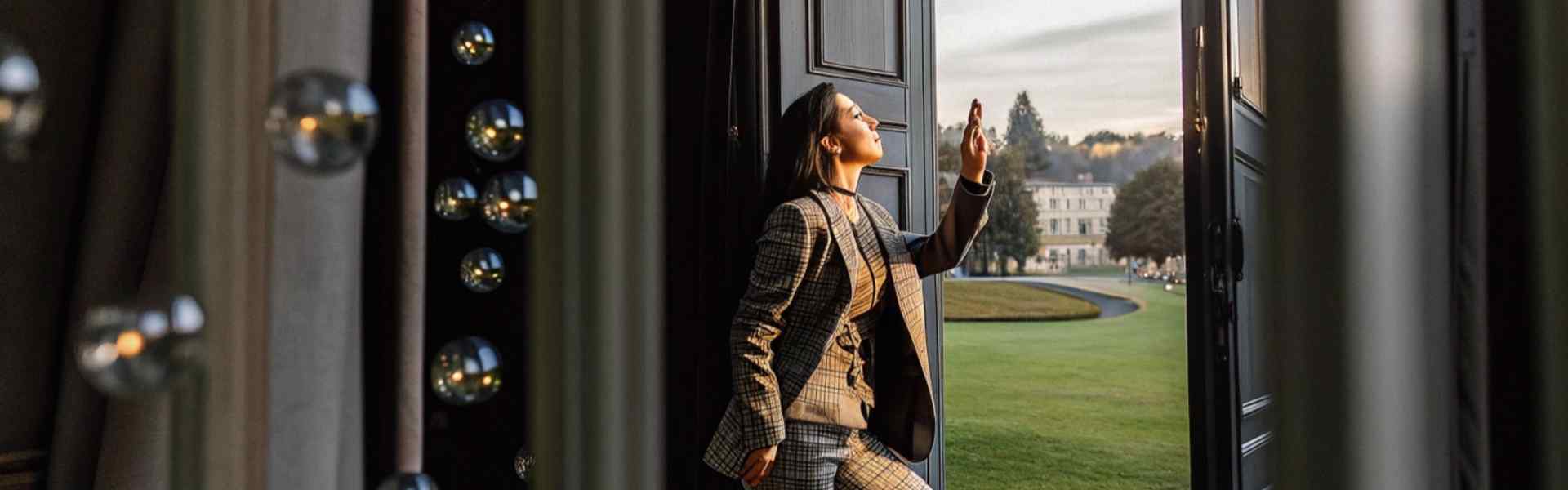 Woman in a plaid suit standing by an open door, gazing at a scenic estate.