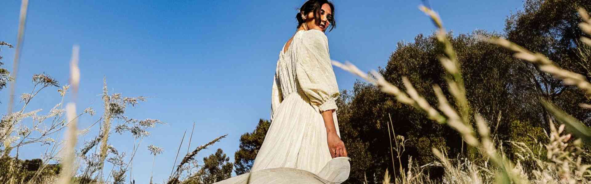 Woman in a flowing white dress walking through a sunlit field under a blue sky.