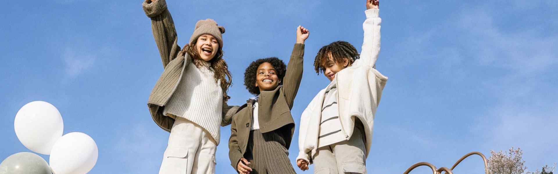 Three children in cozy neutral-toned outfits striking a cheerful pose outdoors.