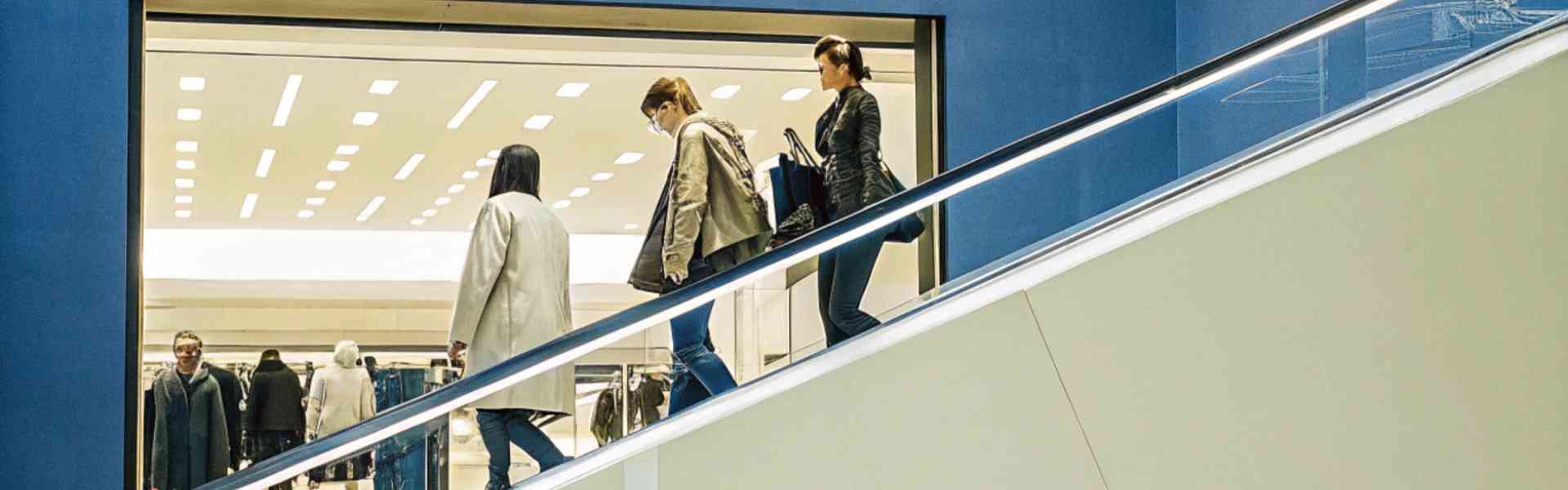 Shoppers descend an escalator in a brightly lit shopping mall with a blue wall.