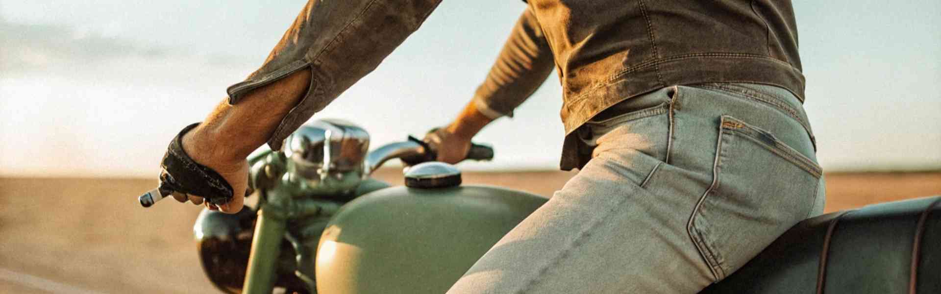 Man wearing distressed jeans riding a vintage green motorcycle in the desert.