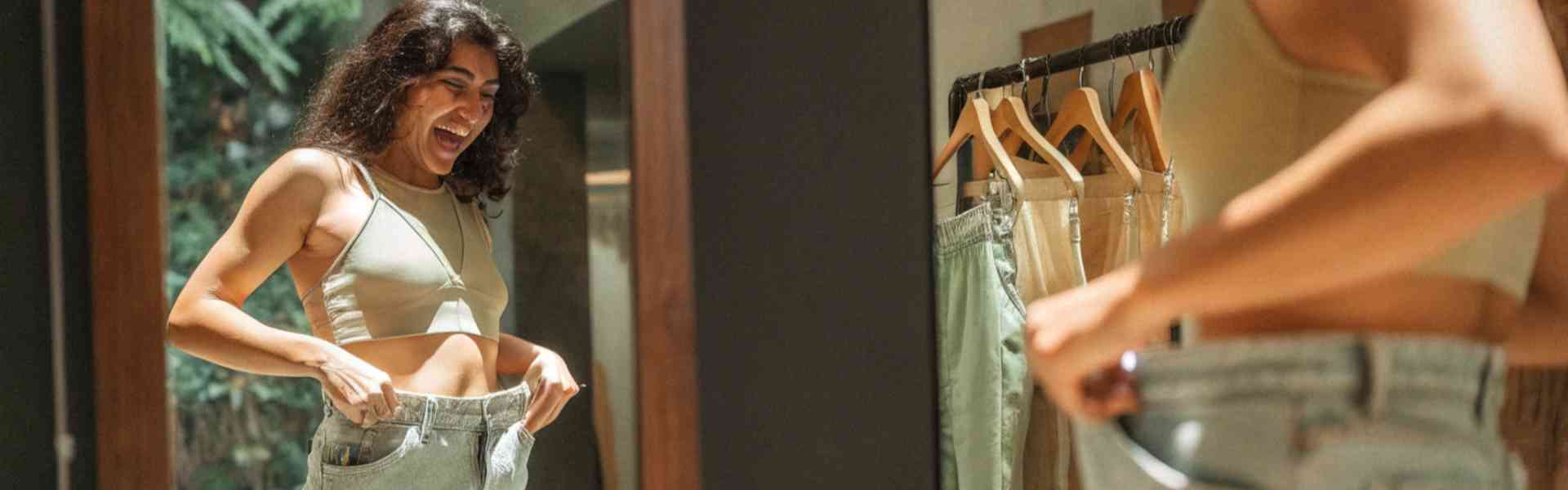 A woman excitedly trying on oversized jeans in front of a mirror.