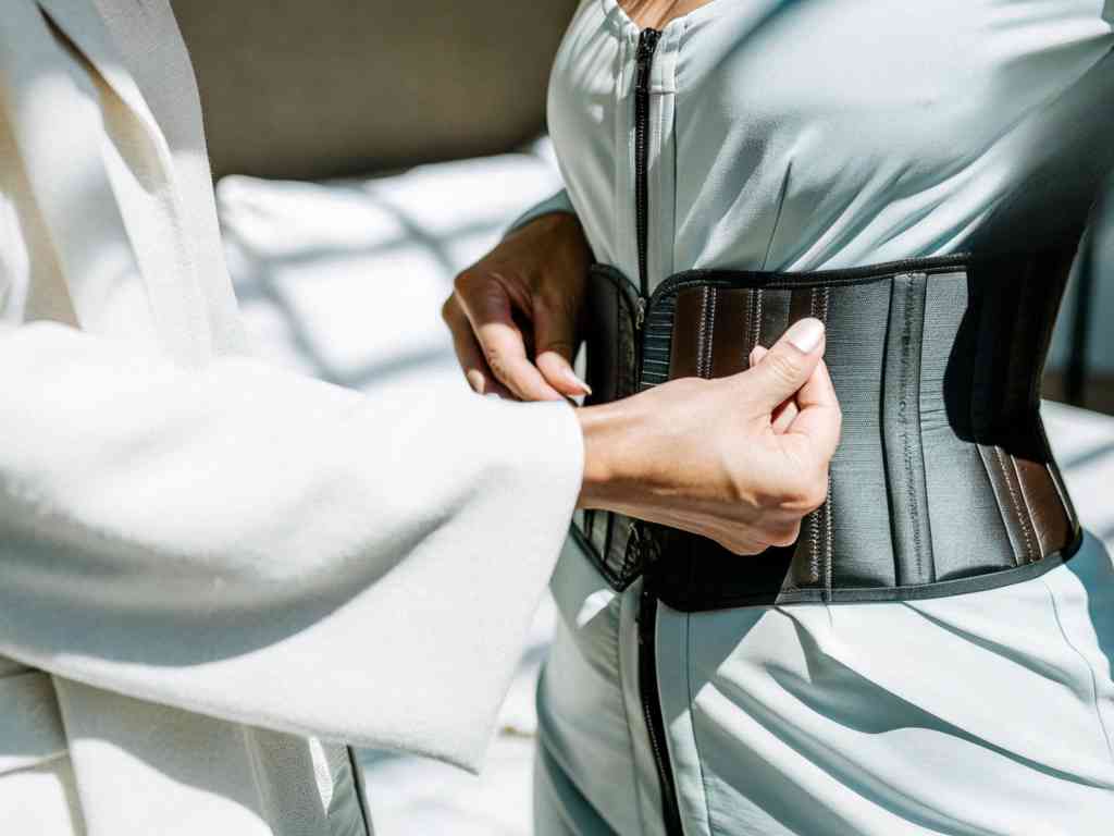 A woman in a light blue outfit tightening a black corset with assistance.