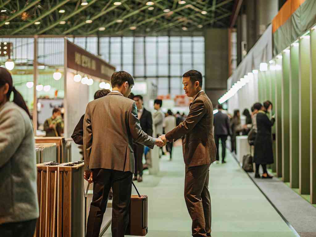 Two businessmen in suits shaking hands at a trade show, surrounded by exhibition booths and other attendees.