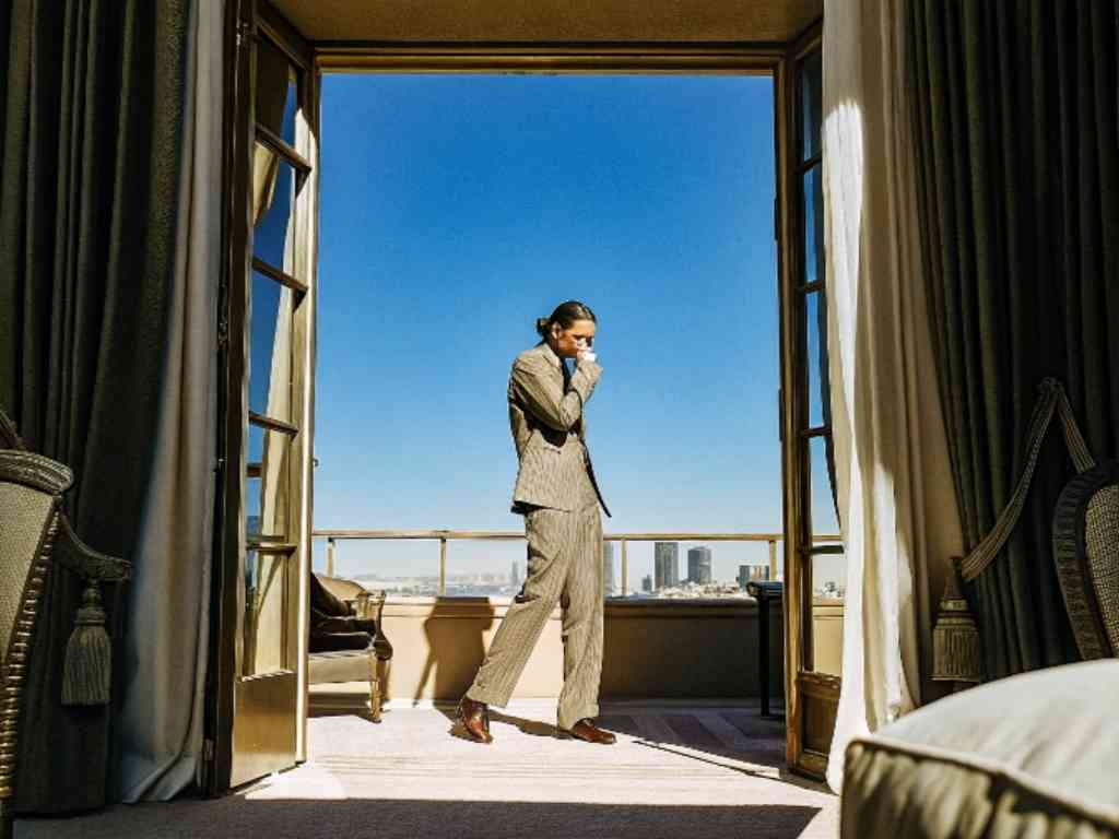 A stylish man in a suit talking on the phone on a balcony with a city view.