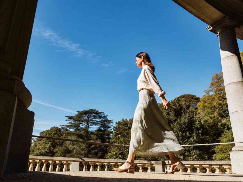 A stylish woman in a pleated skirt and white blouse walks on a terrace under a clear blue sky.