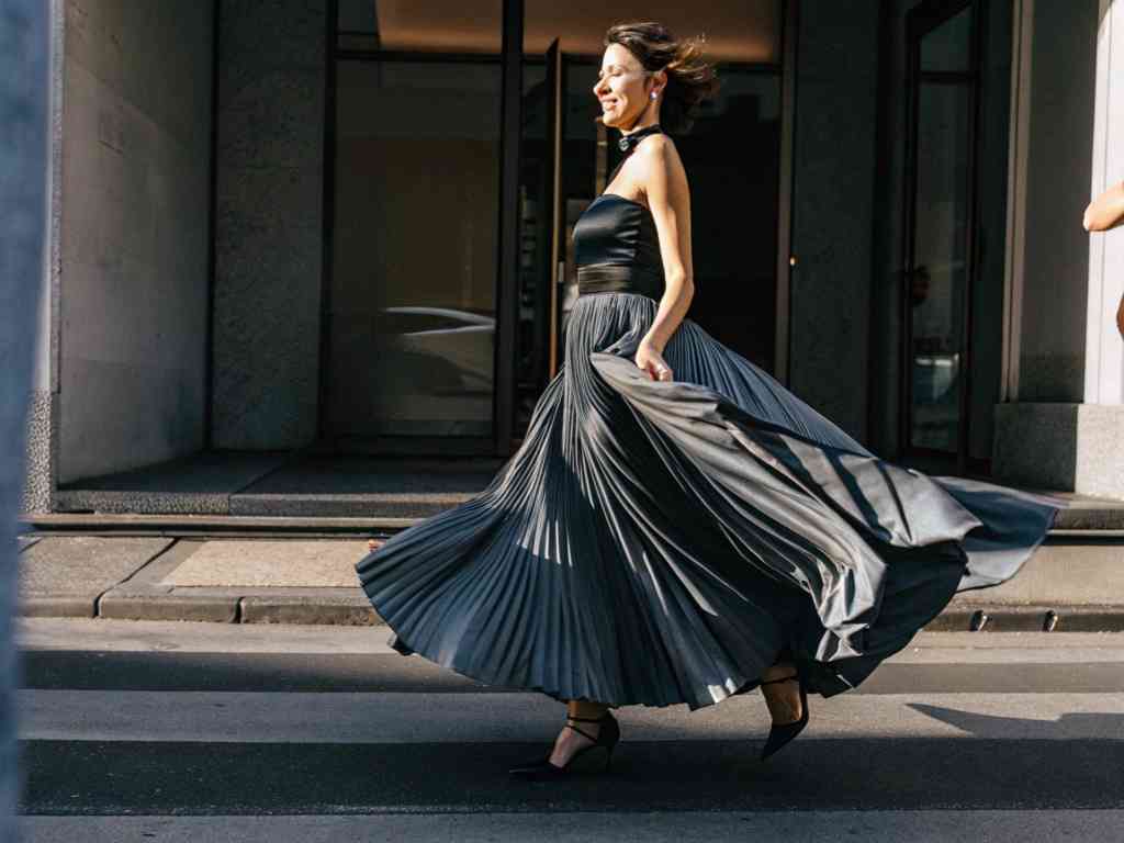 A woman in a flowing black pleated gown joyfully walks across the street, with her dress billowing.