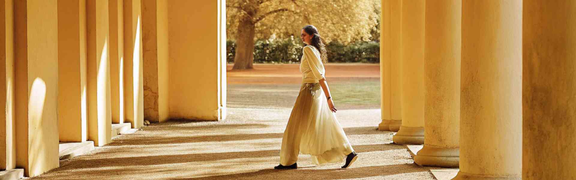 A woman in a flowing white skirt walks through a sunlit, columned walkway with long shadows.