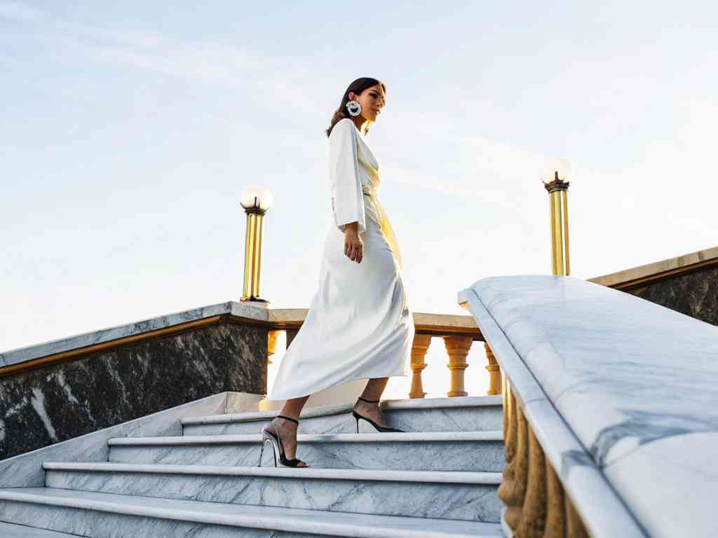 A stylish woman in a flowing white dress walks up marble stairs at sunset.