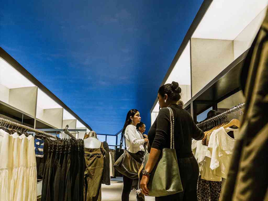 Shoppers browse clothing racks in a modern fashion boutique under bright lighting.