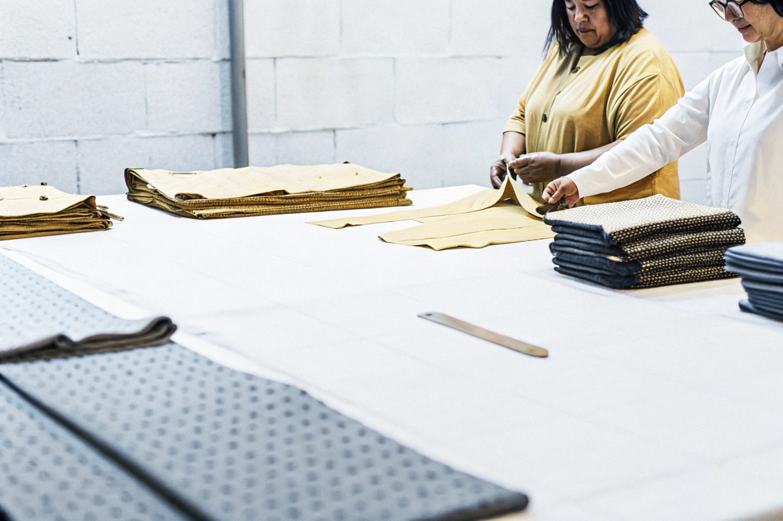 Two women folding and sorting fabric pieces in a workshop with stacks of textiles.