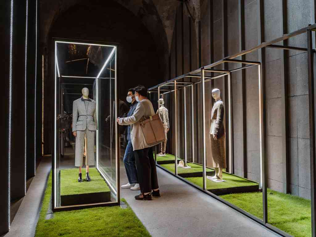 Mannequins in glass cases showcasing stylish outfits at a fashion exhibition, with two people observing.