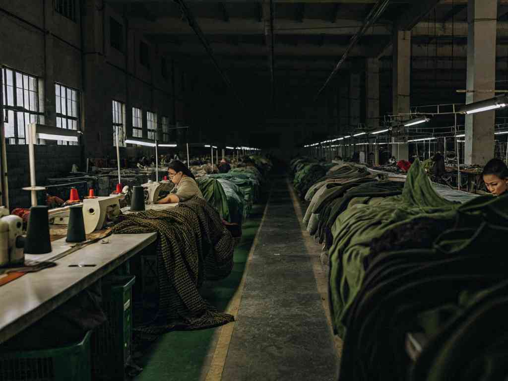 Workers sewing fabric in a dimly lit garment factory with sewing machines and cloth piles.