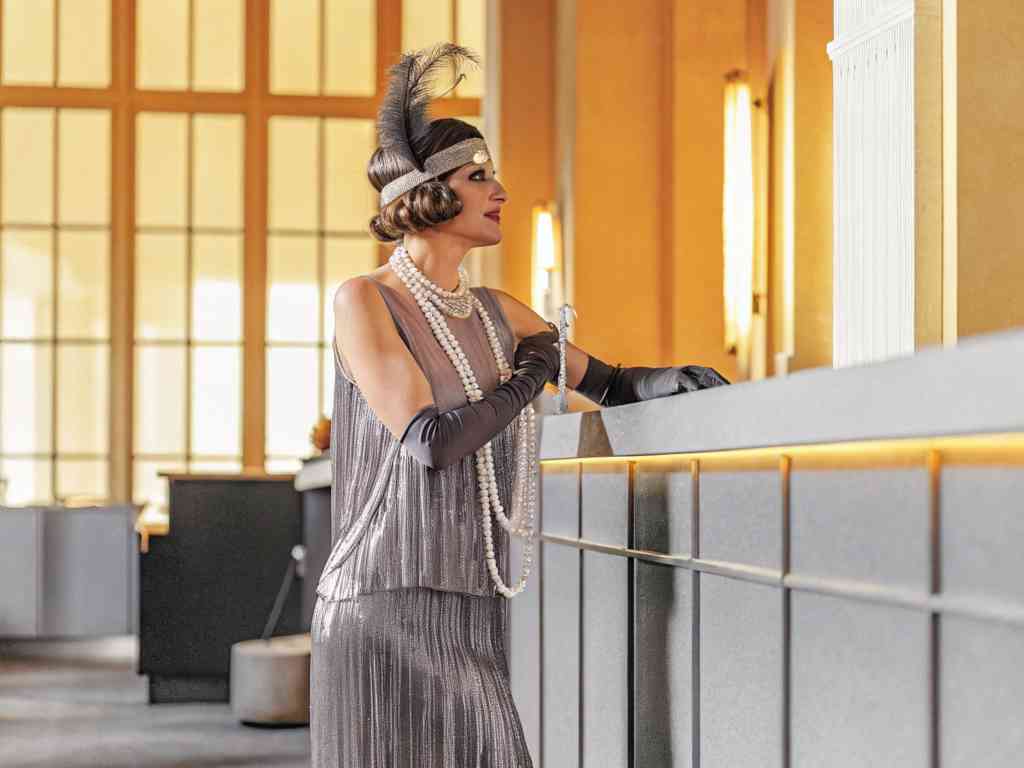 A woman dressed in a silver flapper dress with pearls and gloves in an elegant setting.