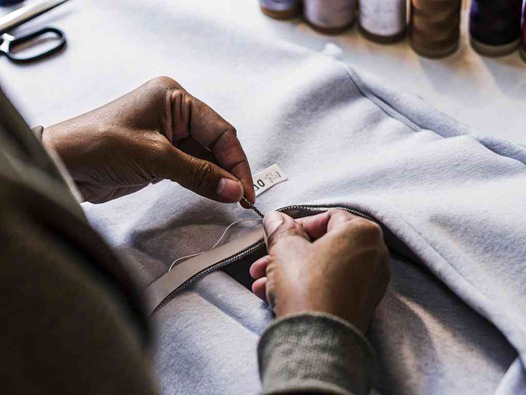 Close-up of hands sewing a label onto a gray garment with thread and needle.