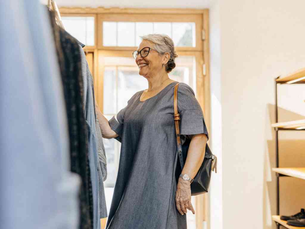 A smiling older woman in a gray dress and glasses browses clothing in a boutique, holding a garment.