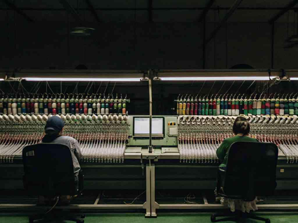 Two workers operating a massive embroidery machine with colorful thread spools.