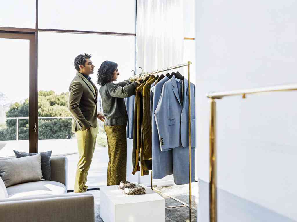 A well-dressed couple browsing suits and jackets on a gold clothing rack.