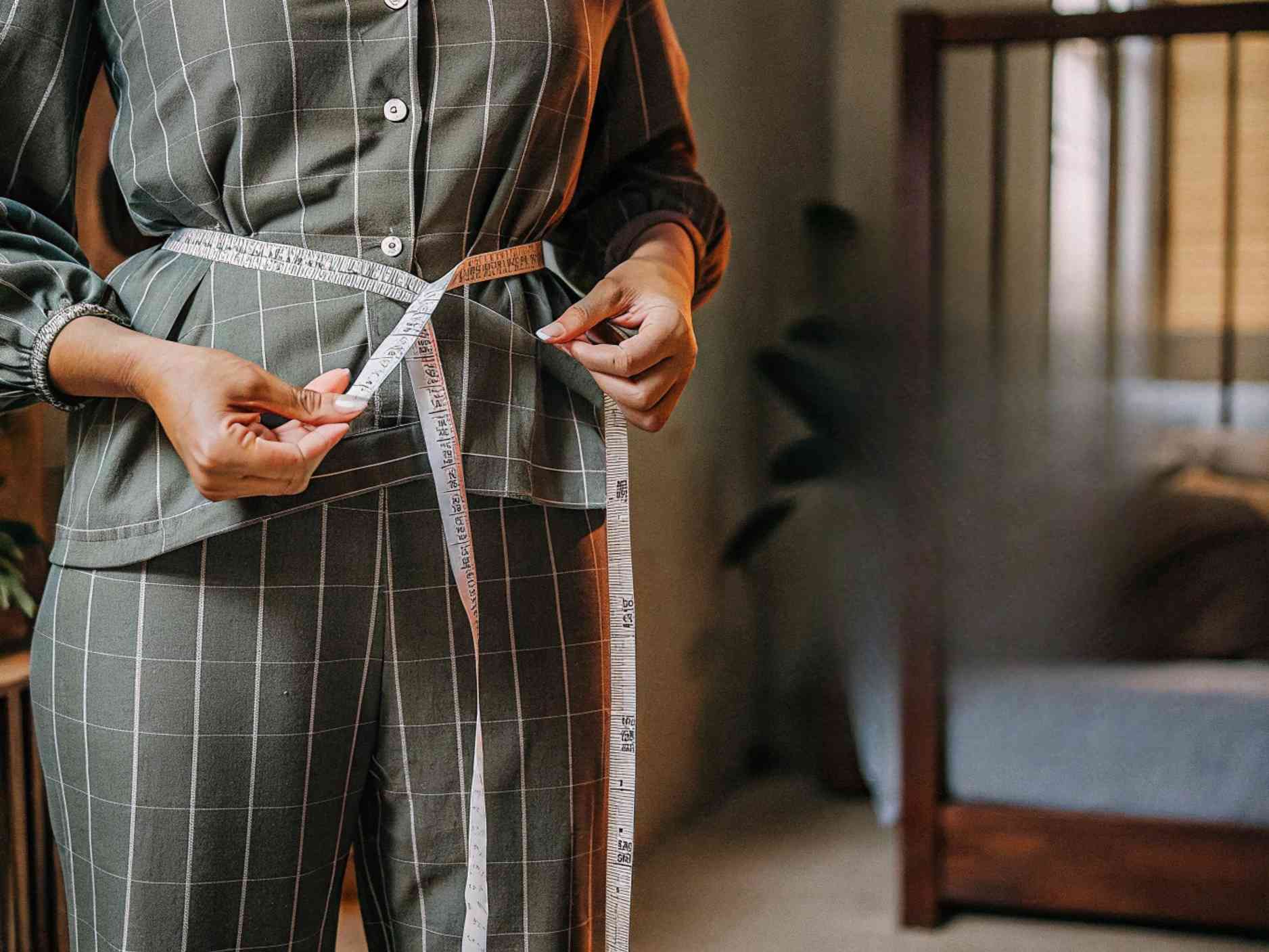 A woman in a checkered outfit measuring her waist with a tape measure.