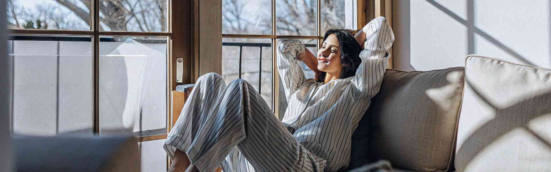 A woman in striped pajamas relaxing on a couch by a large window.