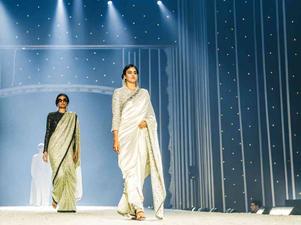 Models in shimmering sarees walk down the runway under bright stage lights.