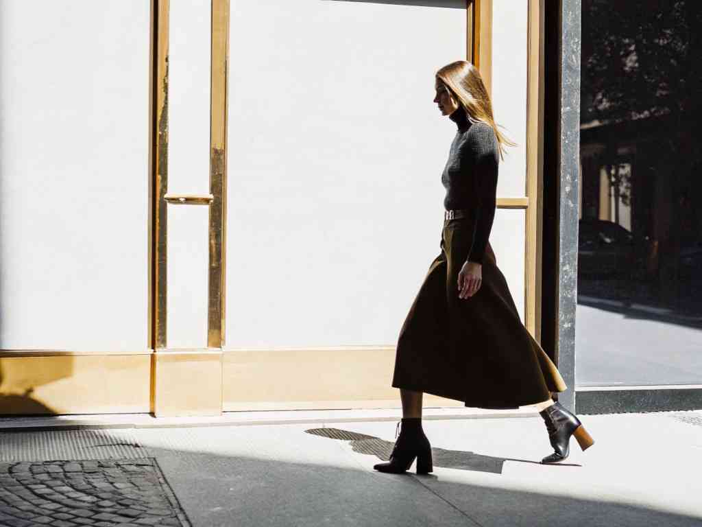 A fashionable woman in a black turtleneck and brown skirt walks past a sunlit wall, casting a shadow.