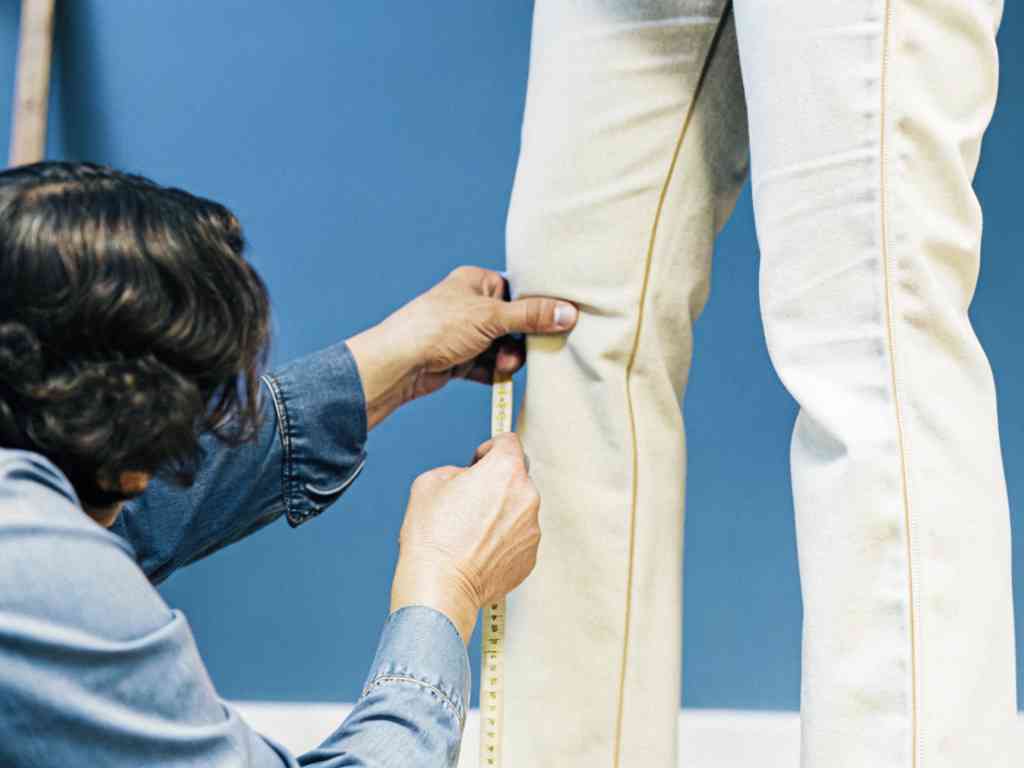 A tailor measures the fit of cream-colored pants on a standing person against a blue background.