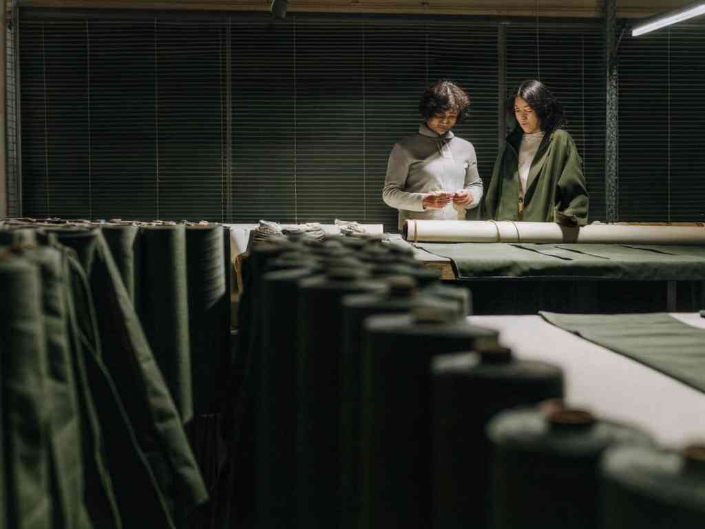 Two women examine fabric rolls in a dimly lit textile workshop with green materials.