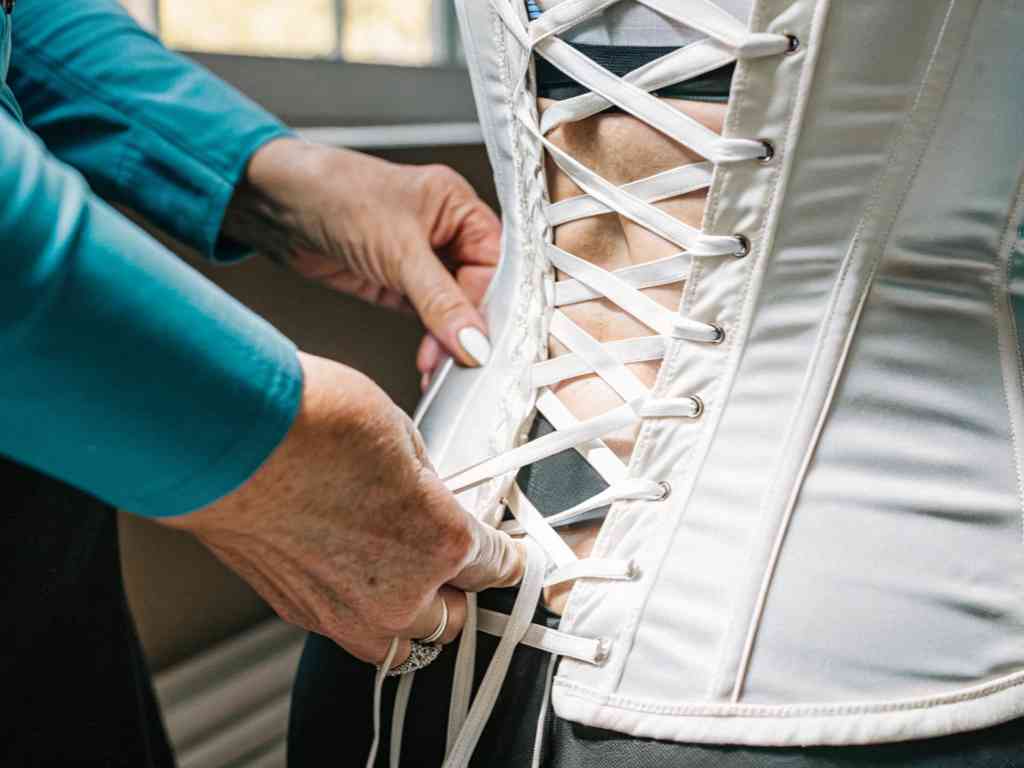 A person tightening the white laces of a satin corset on someone’s back.