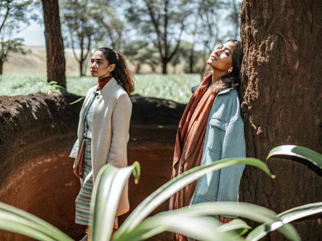 Two stylish women stand in a sunlit forest, one leaning against a tree, both enjoying the natural surroundings.