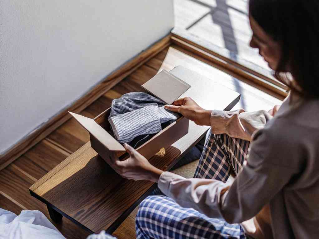 A woman in pajamas opens a small box containing neatly folded clothing in a cozy setting.