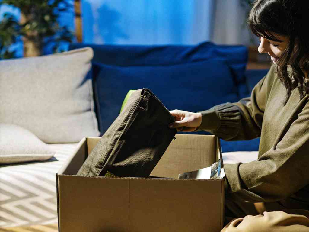 A woman in a cozy setting unpacks a cardboard box, revealing neatly folded clothing.