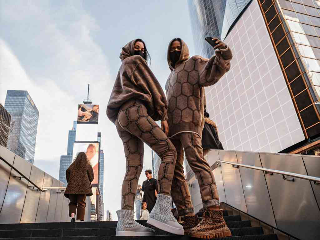Two stylish women in futuristic streetwear take a selfie on a city staircase, surrounded by skyscrapers.