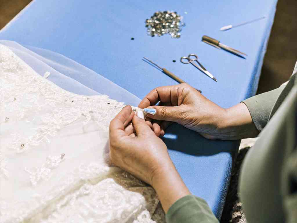 Hands sewing beads onto lace bridal fabric on blue table.