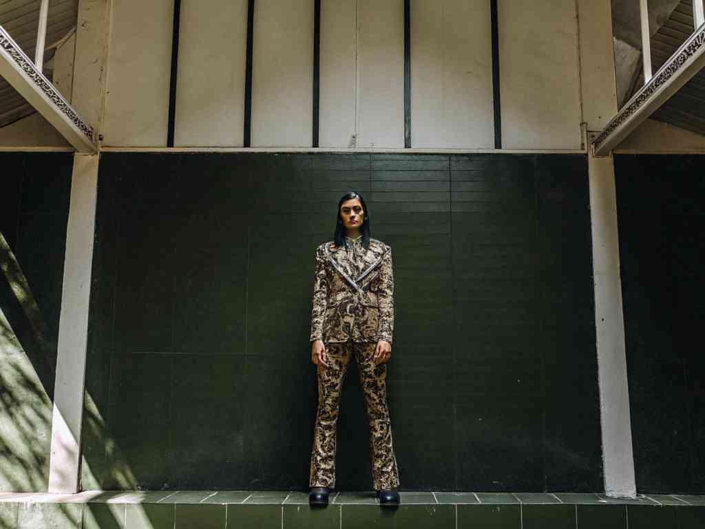 Model in patterned suit poses against green tiled wall