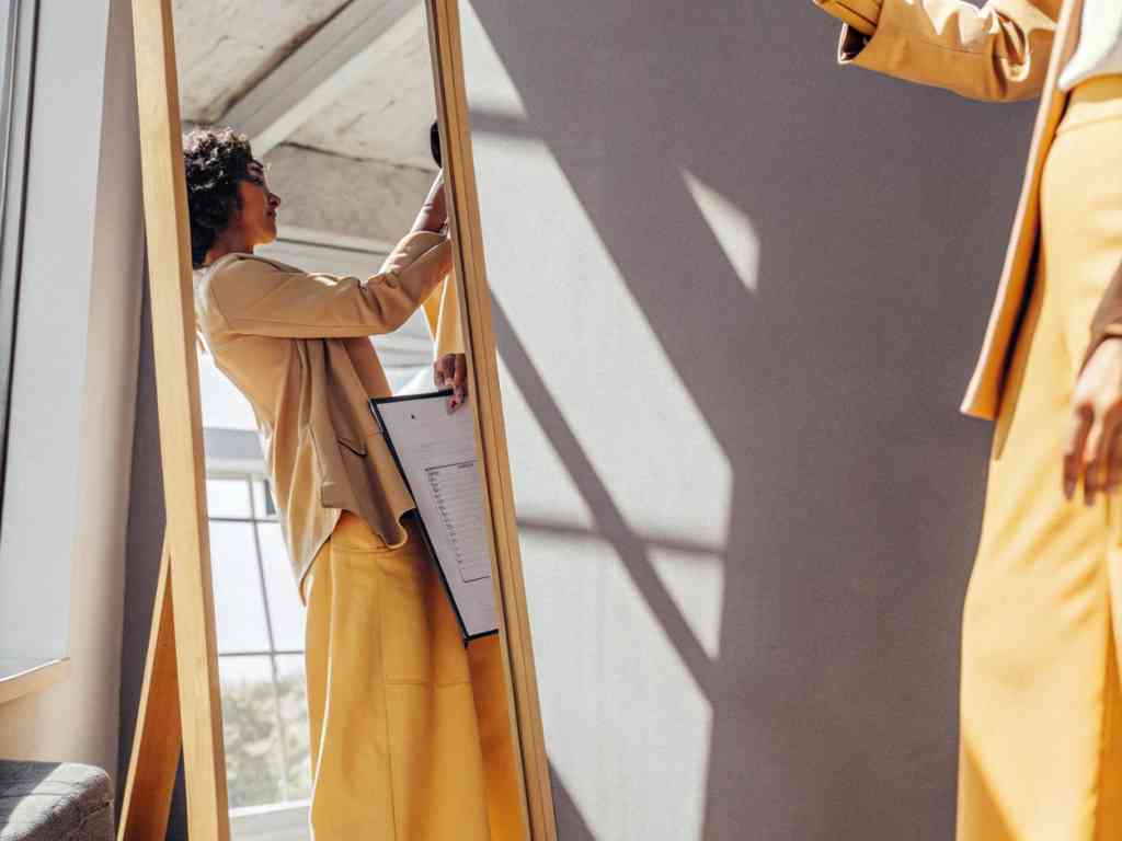 Woman in yellow outfit checking mirror while holding clipboard.