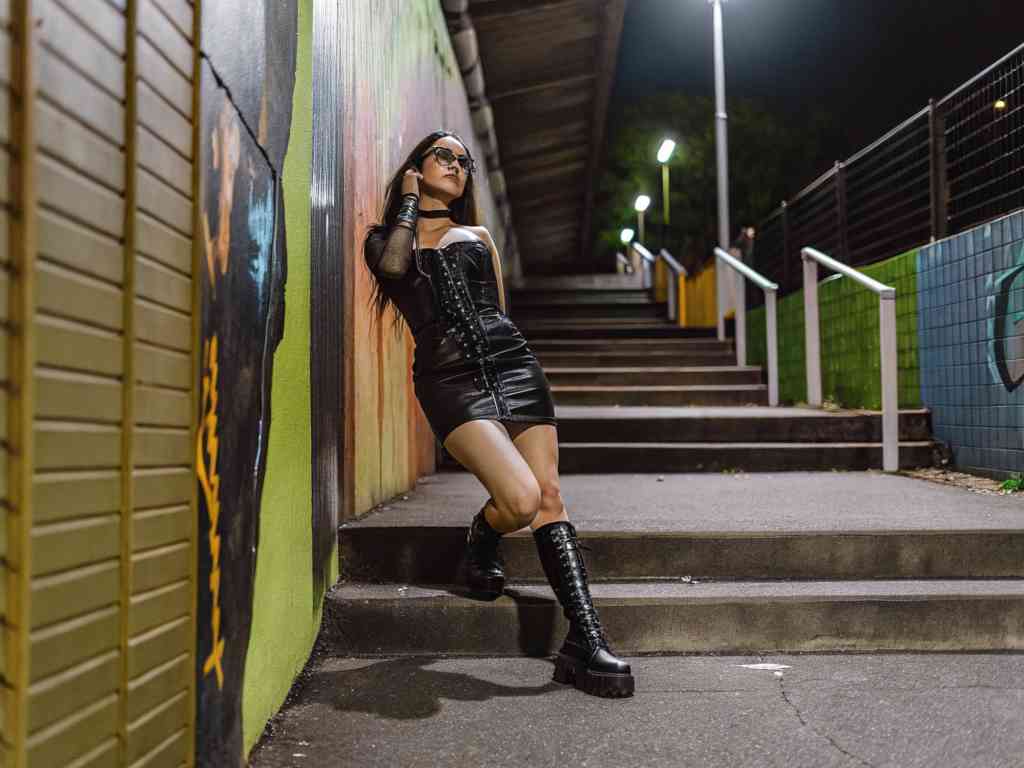 Woman in black leather corset outfit leaning on stairs in graffiti-lit alley.