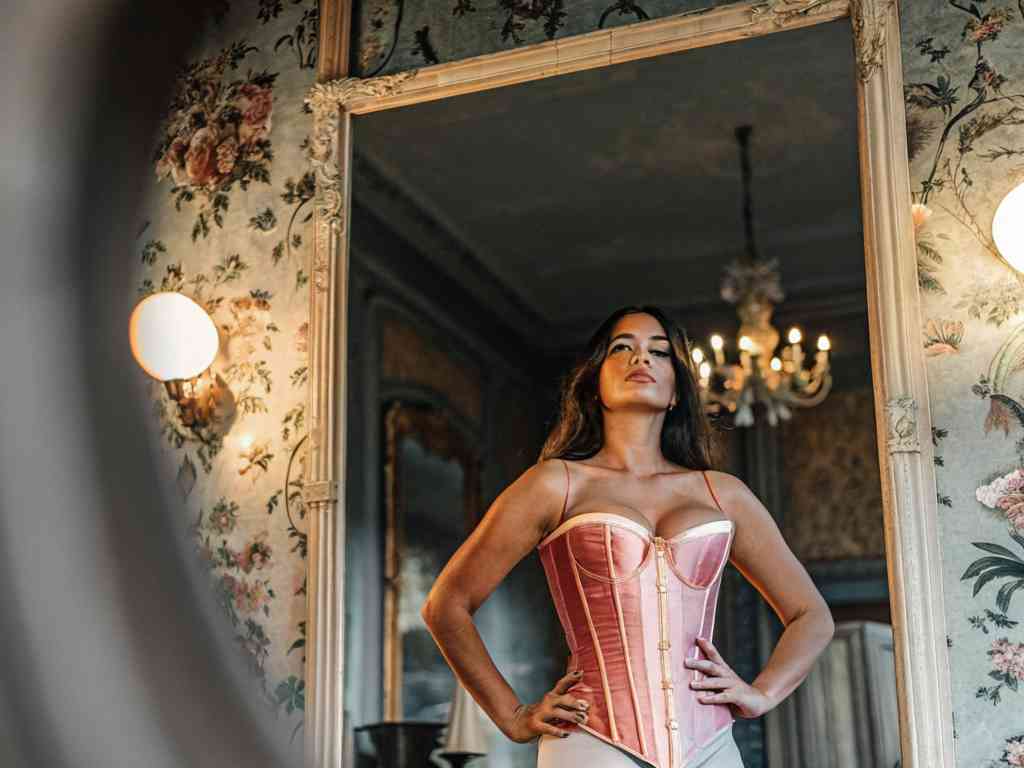 Woman in pink satin corset standing confidently in ornate vintage room.