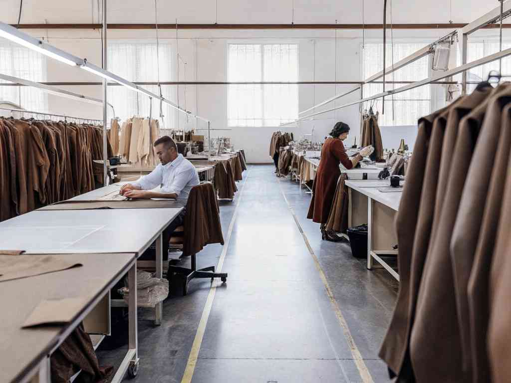 Garment factory workers sewing and preparing brown fabric pieces in a well-lit workspace.