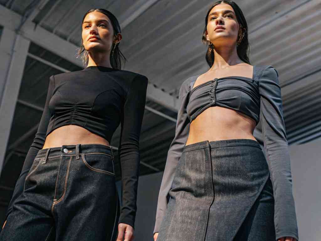 Two models wearing dark denim with fitted crop tops posing under industrial lighting.