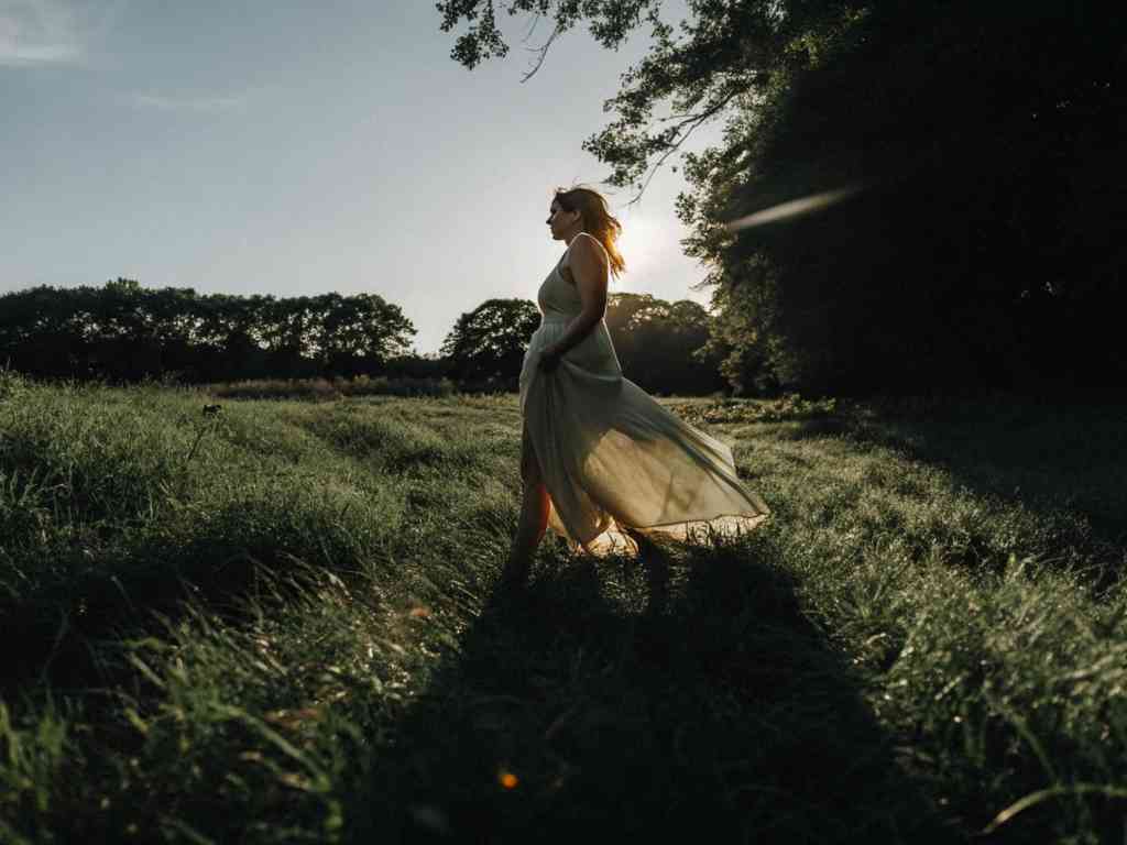 Woman in flowing dress walking through grassy field at sunset.