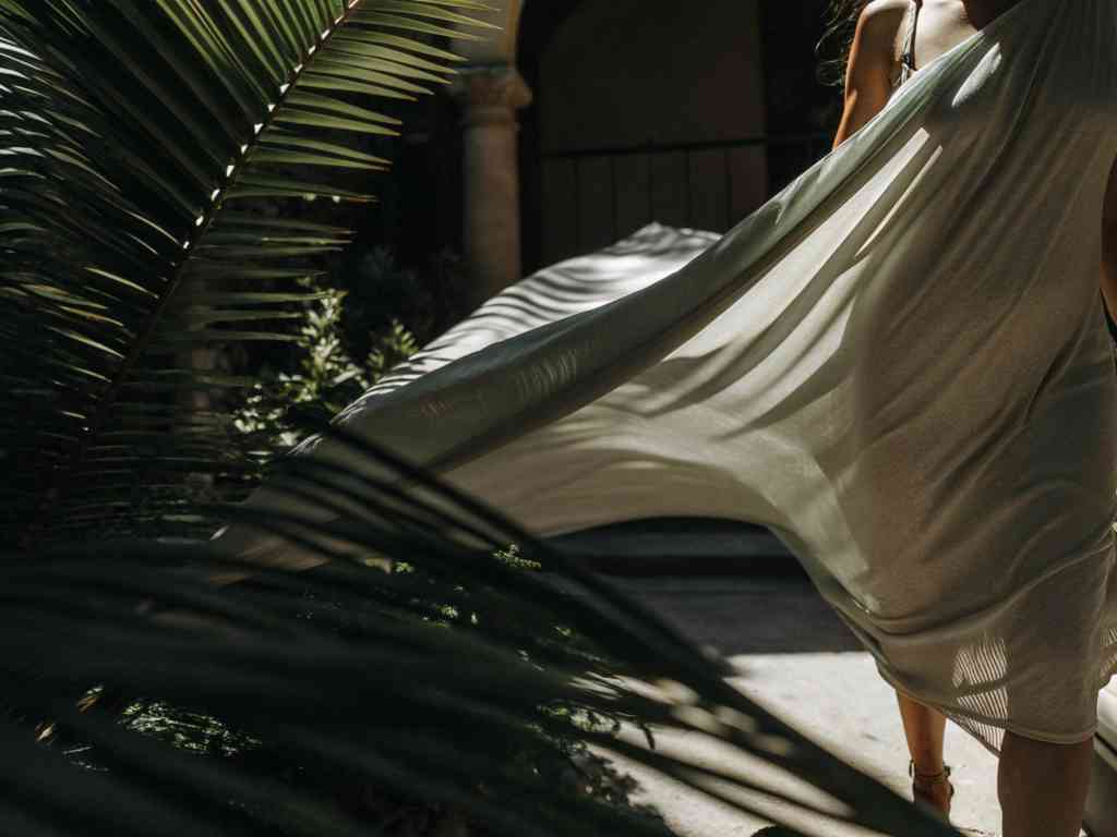 Woman in flowing dress walking past tropical foliage in dappled sunlight.