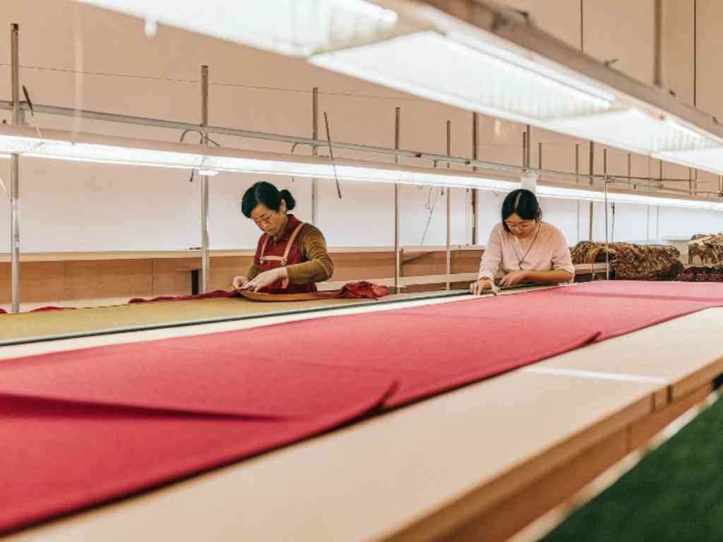 Two women working with red fabric on long garment inspection table.