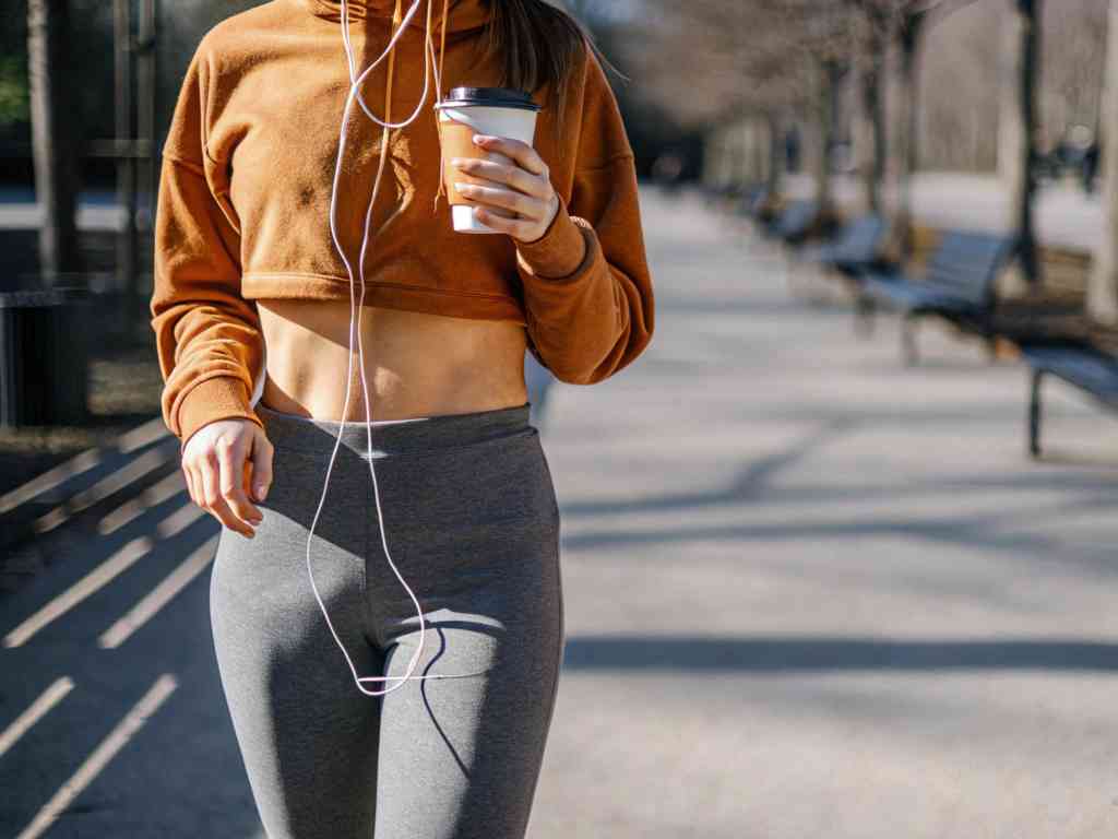 Woman in brown cropped hoodie and grey leggings walking in a park with coffee and headphones.
