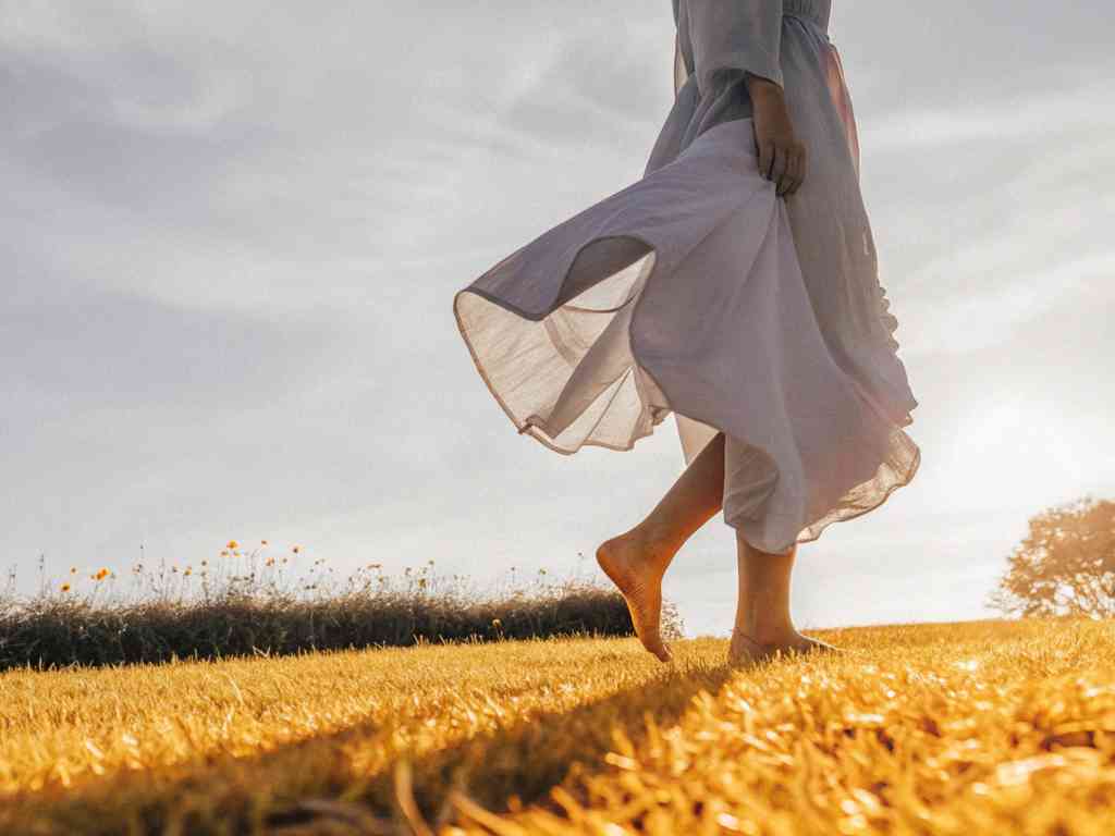 Barefoot woman walking through golden field in flowing dress.