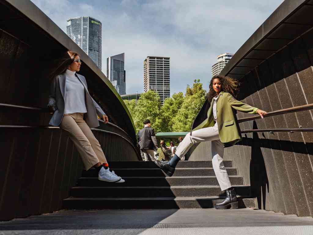Two women in stylish blazers pose on stairs in cityscape