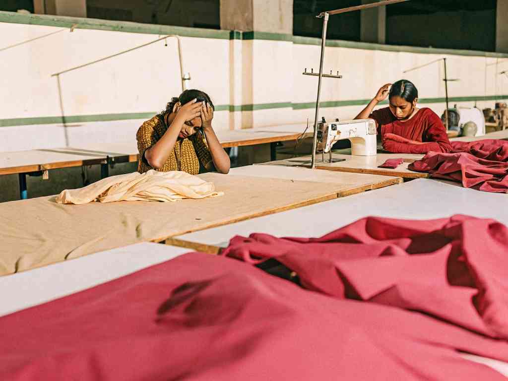Two women working in a garment factory, one appears stressed while the other focuses on sewing fabric.