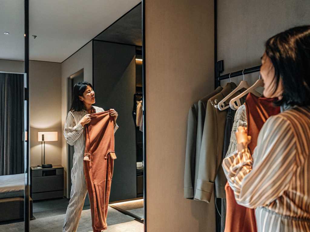 Woman in striped loungewear holding a rust-colored jumpsuit in front of a full-length mirror inside a modern bedroom closet