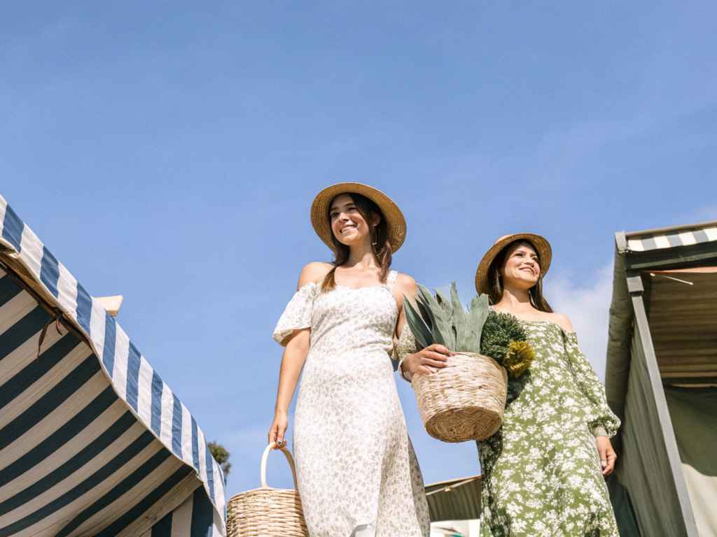 Two women in floral summer dresses and straw hats carrying baskets at an outdoor market.
