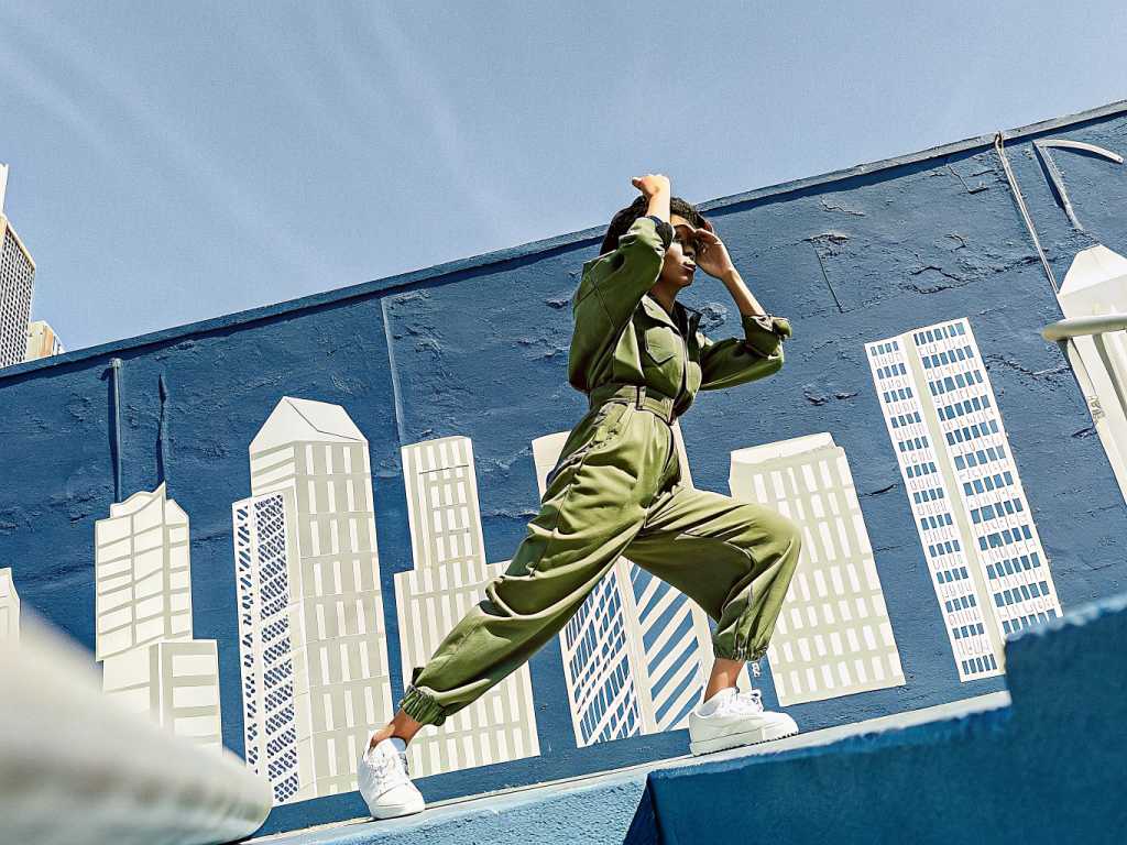 Woman in olive jumpsuit lunges before blue cityscape mural
